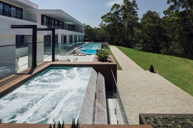 Exterior view of a large contemporary multi-level residence with white rendered walls, extensive dark-framed glazing, and layered flat roof forms. The image focuses on a long outdoor pool terrace with a bubbling spa in the foreground, an additional swimming pool beyond, timber decking, and a broad paved walkway beside landscaped lawn and mature trees.