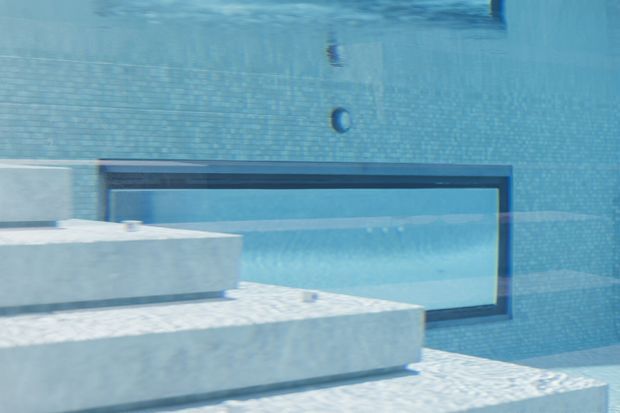 Underwater view of a tiled swimming pool with broad pale concrete or stone steps and a dark-framed rectangular glazed opening or pool feature in the background. The image is dominated by cool blue water and blue mosaic tile surfaces.