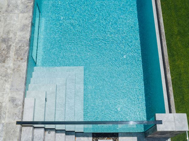 Aerial view of a contemporary rectangular swimming pool with clear turquoise water, broad pale stone steps descending into the pool, and a dark glass or metal railing crossing the foreground. The pool is bordered by light grey stone paving and a narrow strip of lawn.