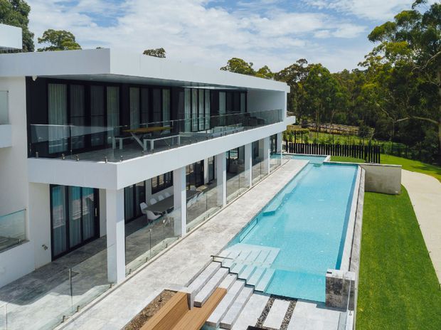 A large contemporary white multi-storey residence with extensive floor-to-ceiling glazing, dark-framed windows, wraparound balconies, and transparent glass balustrades. A long rectangular swimming pool runs alongside the house, with broad pale stone paving, stepped entry, timber decking, and manicured lawn. The property is set among mature trees under a bright sky.