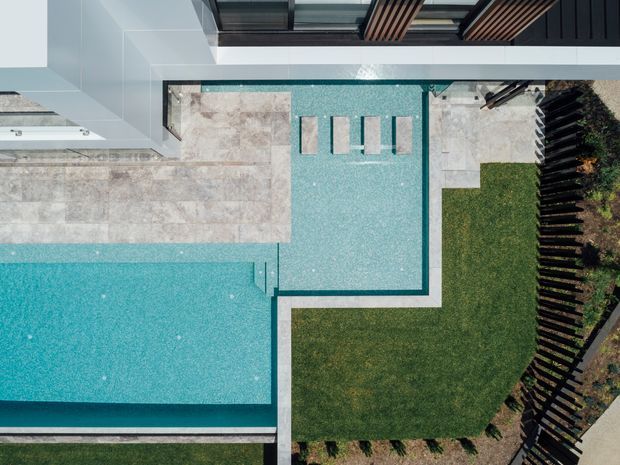 Aerial view of a contemporary residential outdoor area with two connected turquoise swimming pools, pale stone paving, a neatly edged lawn, and dark slatted fencing. Modern building edges, glazing, and timber-like overhead screening are visible along the upper edge of the composition.