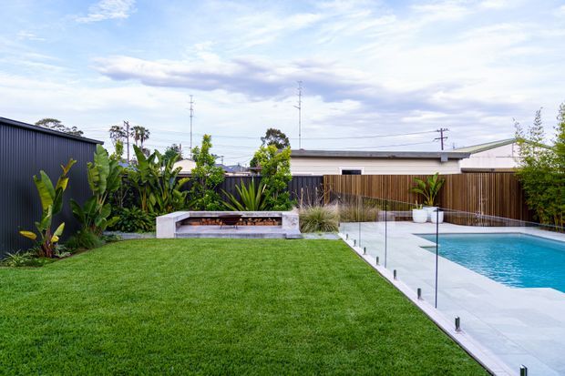 A landscaped backyard with a broad green lawn, a rectangular swimming pool bordered by frameless glass safety fencing, and a pale paved pool surround. A low rectangular outdoor fireplace or water feature sits among tropical planting and ornamental grasses, with timber and dark metal boundary fencing and a single-storey residential building beyond. The scene is bright, contemporary, and carefully designed.