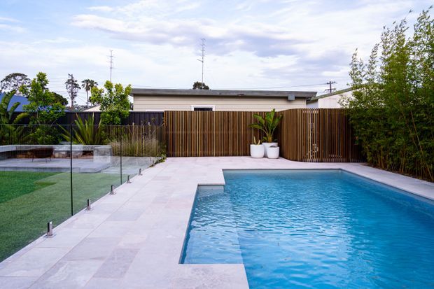 Contemporary residential backyard with a rectangular swimming pool surrounded by pale stone or concrete paving. A timber slat privacy fence, tropical planters, glass pool fencing, landscaped lawn and a low flat-roofed structure create a clean, private outdoor entertaining space.