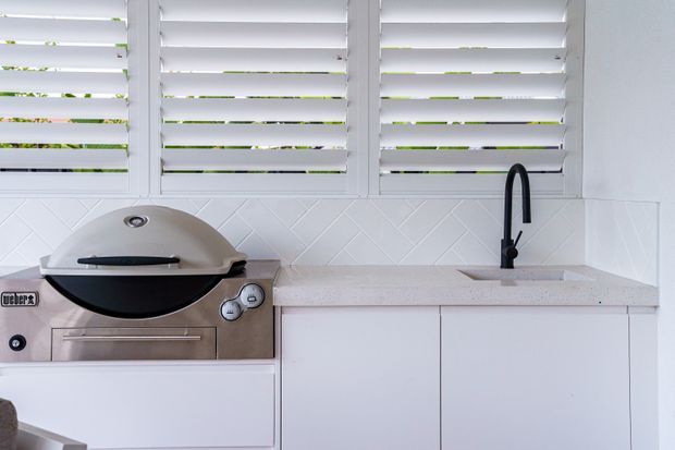 A compact white outdoor kitchen with built-in cabinetry, a pale stone benchtop, a built-in barbecue, black gooseneck tap and sink, white geometric tiled splashback, and white louvred window shutters. The space is bright and clean with a restrained contemporary coastal feel.