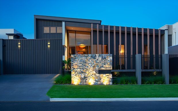 Contemporary single-storey residential facade at dusk, featuring dark vertical cladding, a recessed warm-lit entry, slim vertical screens, and a prominent illuminated natural-stone feature wall. The property fronts a paved driveway and neatly landscaped lawn.