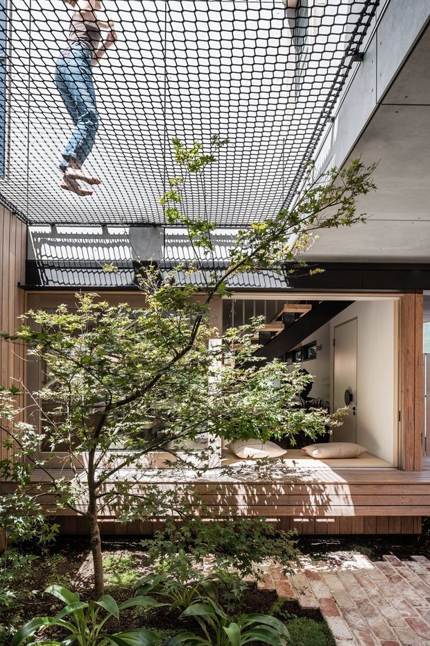 A bright, compact courtyard-style residential space with a timber-clad raised deck, sliding glazed openings, and an open interior beneath a suspended rope-net play area. Japanese maple trees and lush planting fill the foreground courtyard, while a small loft stair and minimalist interior are visible through the opening. Strong sunlight creates patterned shadows across the deck and walls.