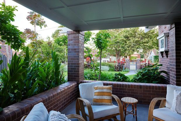 Covered brick patio or veranda with built-in brick planter walls and square brick columns. The space contains woven lounge chairs, cushions, a small side table, and dense tropical planting, opening toward a landscaped residential frontage.