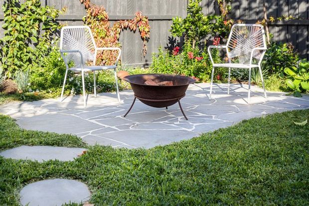 A sunny landscaped garden courtyard with an irregular grey stone-paved sitting area, two white woven outdoor chairs, and a central rusted metal fire pit. Planting borders the space, with a dark vertical-clad wall forming the backdrop and a lawn with stepping stones in the foreground.