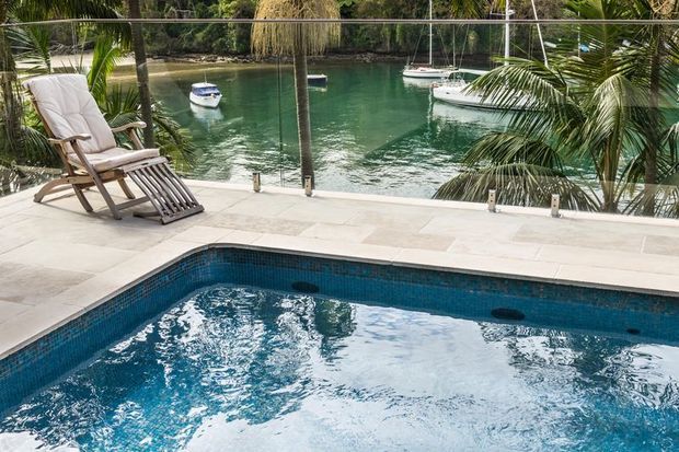 A tropical waterfront pool terrace overlooking a calm canal or marina. The foreground features a rectangular swimming pool with blue mosaic tile lining, pale stone paving, glass balustrading, and a timber reclining lounge chair. Palm trees and moored boats are visible beyond the glass edge.