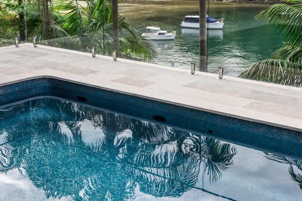 A tropical waterfront swimming pool with dark blue mosaic tile lining and pale stone coping, overlooking calm green-blue water with boats and dense palm vegetation beyond. The pool occupies the foreground, creating strong reflections of the surrounding palms.