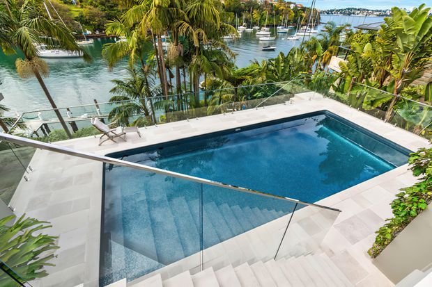 Elevated view of a modern outdoor swimming pool terrace with broad pale stone paving, glass balustrades, and wide staircases descending into the pool area. Tropical planting surrounds the terrace, with a marina and boats visible beyond.
