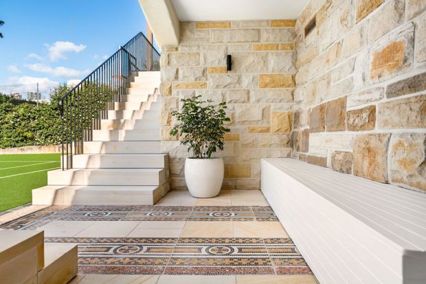 Bright outdoor terrace or poolside entertaining area beside a rendered and natural stone-clad wall. A broad tiled stairway with black metal railing leads upward, while patterned decorative floor tiles, a long white built-in bench, potted greenery and an adjacent artificial-turf sports court define the space.