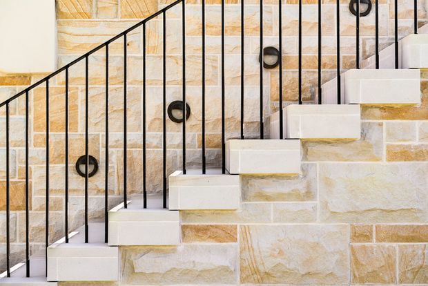 Close-up view of a contemporary staircase with white solid treads and risers set against a textured natural stone masonry wall. A slim black metal balustrade with vertical rods and circular detail elements runs alongside the stair, creating a strong geometric contrast.