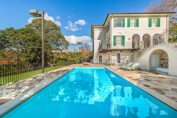 A sunny landscaped swimming pool courtyard beside a large white rendered multi-storey residence. The house features green shutters, arched windows and openings, a curved exterior staircase, wrought-style balustrades, and a stone-paved pool terrace. Mature trees and distant city buildings are visible beyond the property.