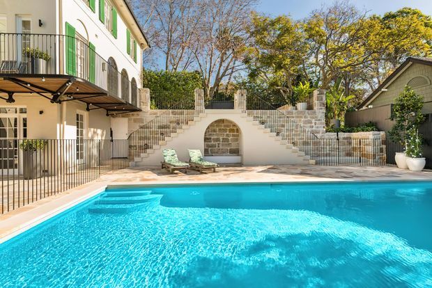 Bright exterior view of a landscaped residential pool courtyard. A large turquoise swimming pool occupies the foreground, bordered by pale paving and a low wall with a central arched stone feature. Symmetrical stairs rise to a planted upper terrace, while the surrounding cream-rendered architecture has dark metal balcony railings, green shutters, and stone detailing. A small pitched-roof outbuilding is visible to the right.