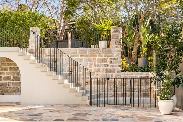A landscaped exterior courtyard or terrace featuring a rendered stairway with slim black metal balustrades, natural stone retaining walls and columns, and patterned natural-stone paving. Mature trees and tropical planting surround the space, creating a refined garden setting.
