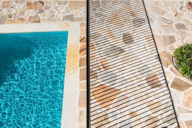 Overhead close-up of a bright turquoise swimming pool bordered by pale irregular natural-stone paving and a narrow timber-toned strip. A black metal-framed slatted outdoor lounge or poolside structure occupies the right side, with ornamental planting at the edge.