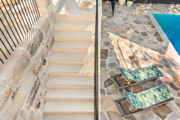 Outdoor stone-paved pool area viewed from a stairway, with pale stone steps and retaining walls, black metal railings, patterned natural-stone paving, and a bright blue swimming pool. Two sun loungers sit beside the pool.