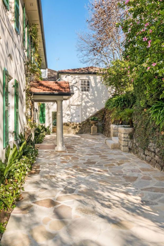 A sunlit landscaped courtyard and external walkway between white rendered residential buildings. The path is paved with irregular natural stone, bordered by lush planting and a low stone retaining wall. Green louvred shutters, terracotta tiled roofs, and a covered entry supported by a classical column contribute to a Mediterranean traditional character.