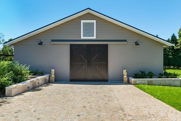A symmetrical single-storey gable-roof building with pale horizontal siding, dark timber double barn doors, and a central louvred vent. A broad irregular stone-paved driveway leads to the entrance, flanked by low stone retaining walls, planting, and small urn-like ornaments.