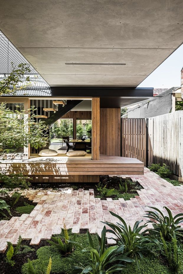 A contemporary courtyard garden viewed from outside, featuring a compact timber-clad pavilion or extension with a broad exposed concrete roof canopy, large glazed opening, and built-in timber bench/deck. The interior reveals a dining area and open stair. Pale reclaimed brick paving, raised planted garden beds, vertical timber fencing, and layered greenery create a strong indoor-outdoor connection.