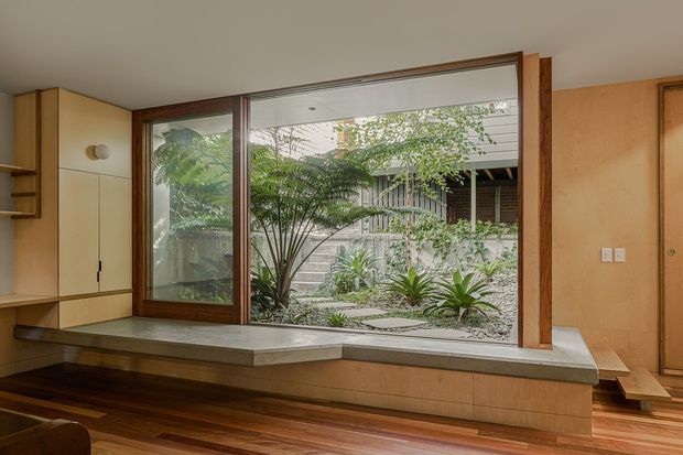 Warm contemporary interior centered on a large timber-framed glazed opening overlooking a landscaped courtyard. Built-in pale wood cabinetry and a long stone-topped window seat line the wall, while dark timber flooring and a wood-panelled doorway add richness. The courtyard features layered planting, gravel, and a screened neighbouring structure visible through the glazing.