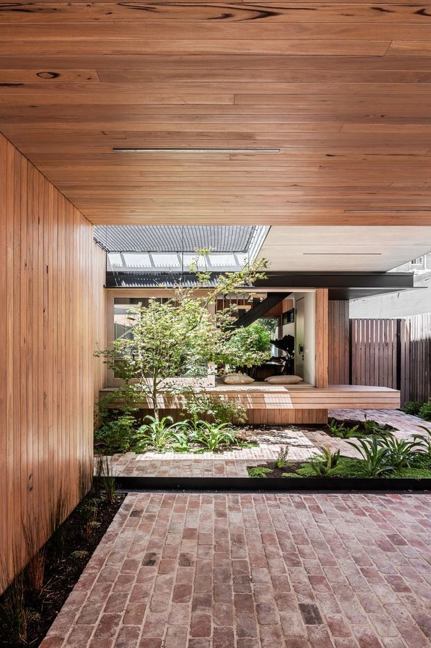 A contemporary courtyard viewed from beneath a warm timber-lined canopy. The space features vertical timber wall cladding, exposed timber soffit, red-brown brick paving, raised garden beds with lush planting, and a large opening connecting to an interior living area. Strong geometric lines, layered indoor-outdoor transitions, and filtered daylight define the composition.