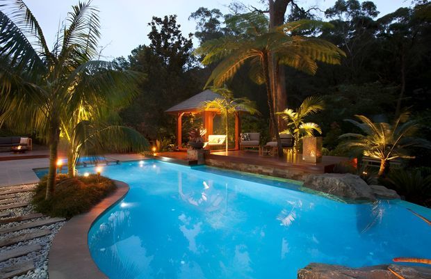 A landscaped outdoor pool area at dusk, featuring a large curving turquoise swimming pool surrounded by tropical palms, dense greenery, natural stone edging, and a warmly lit timber pavilion with outdoor seating. The scene has a resort-like, tranquil atmosphere.