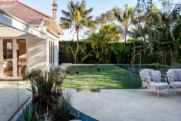 A bright tropical courtyard and poolside outdoor living area beside a light-painted residential building. The scene features a pale stone-paved terrace, a small curved pool or water feature with blue mosaic tiling, white outdoor lounge chairs, dense tropical planting, and a traditional terracotta tiled roof with classical trim and chimney.