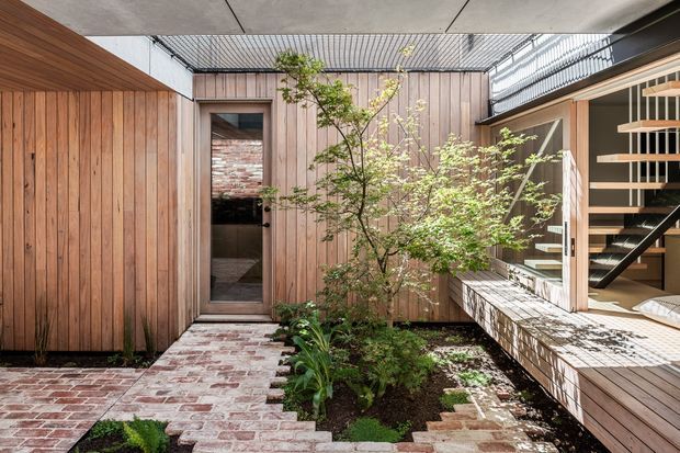 A modern enclosed courtyard with warm vertical timber-clad walls, a red brick pathway and stepped paving, integrated planting, and an open glazed area revealing a timber staircase. The courtyard is partially covered by a concrete slab and screened overhead, creating a private indoor-outdoor connection.