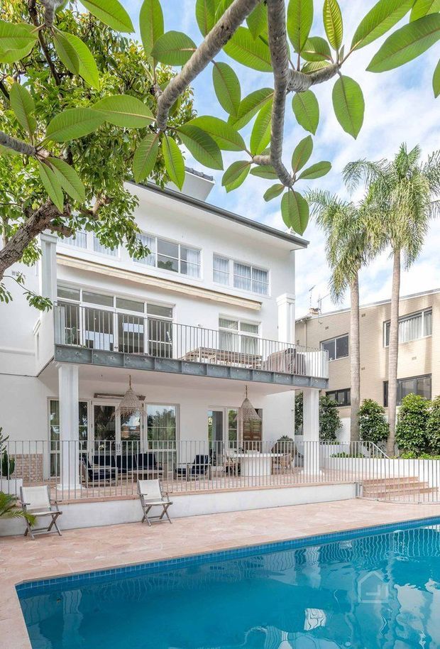 Bright exterior view of a multi-storey white rendered residence beside a swimming pool. The house features broad balconies and terraces with white metal railings, large glazed doors and windows, slender columns, and a covered outdoor living area. Tropical planting and palm trees frame the scene, with terracotta-toned paving around the pool.