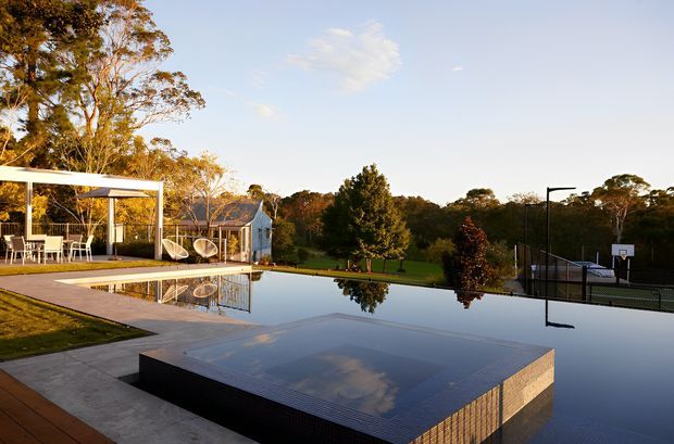 A landscaped residential outdoor area featuring a large reflective swimming pool, a raised tiled pool platform, and a covered outdoor entertaining pavilion with dining furniture and a hanging swing. Mature trees and lawn surround the pool, with a fenced basketball court visible in the background.