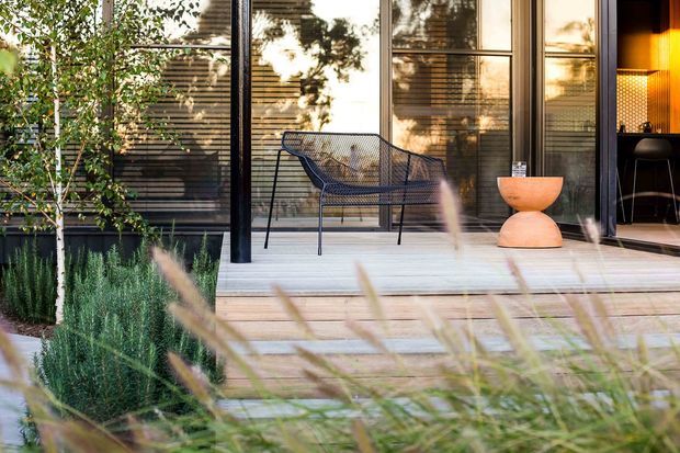 A contemporary outdoor deck and covered terrace viewed from the garden, with pale timber decking, a black wire-frame lounge chair, and a sculptural orange side table. Full-height glazed doors open into a warmly lit interior, while layered grasses and planting frame the foreground.
