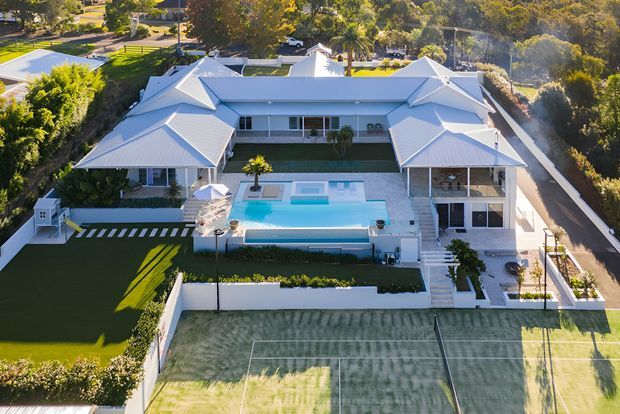 Aerial view of a large, symmetrical light-coloured residential property with multiple pitched roof forms, broad verandas, a central swimming pool and landscaped grounds. A tennis court occupies the foreground, with mature trees and neighbouring properties surrounding the site.