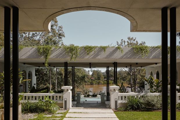 A formal covered outdoor pavilion or terrace frames a landscaped garden and swimming pool, with a small pond or water feature beyond. The structure has broad flat concrete roof slabs, slender black columns, white rendered balustrades and classical arched openings, creating a refined indoor-outdoor connection. Tropical and subtropical planting surrounds a central paved path.