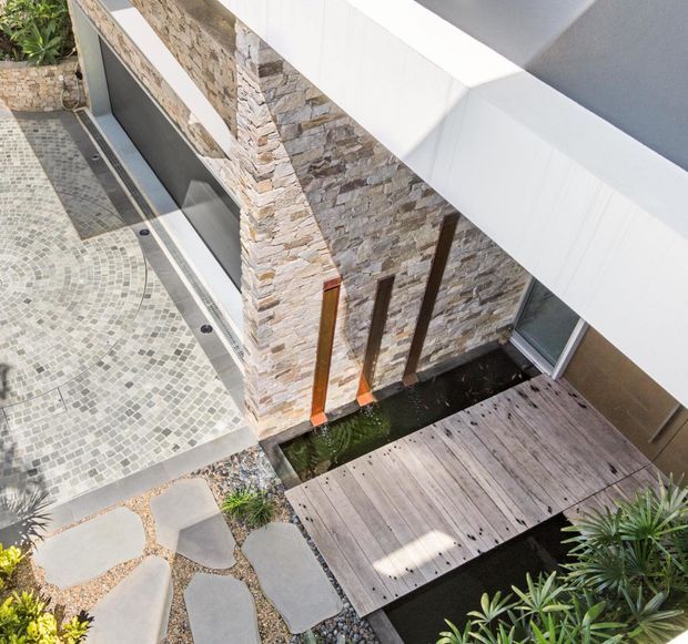 Elevated view of a modern residential exterior courtyard featuring a white angular roof or canopy, textured stacked-stone feature walls, timber vertical accents, a weathered timber deck bridge over a narrow water feature, and paved pathways with mosaic-style stone surfacing and tropical planting.