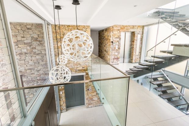 Bright contemporary multi-level interior featuring a double-height circulation space with exposed rough stone walls, extensive glazing, glass balustrades, floating dark stair treads, and sculptural white pendant lights. A lower-level entry or hallway is visible below, with an additional stair and lift or door area in the background.