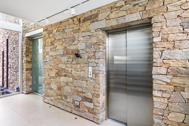 Interior lift lobby or hallway with a prominent rough-stacked natural stone feature wall, stainless-steel elevator doors, a glazed doorway, pale large-format tiled flooring, and recessed track lighting. The space has a contemporary architectural fit-out with strong natural-material detailing.