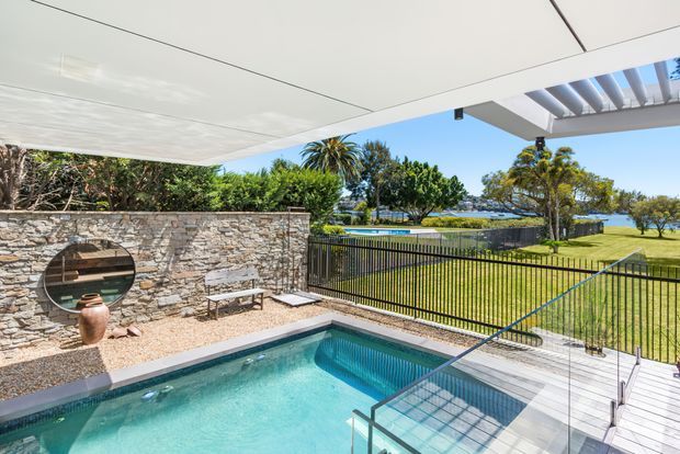 A bright outdoor pool terrace beneath a contemporary covered structure, with a turquoise swimming pool, glass balustrade, pale timber decking, stone feature wall, built-in bench seating, and landscaped lawn beyond. The waterfront is visible in the background, with a pergola-like roof detail and mature trees framing the scene.
