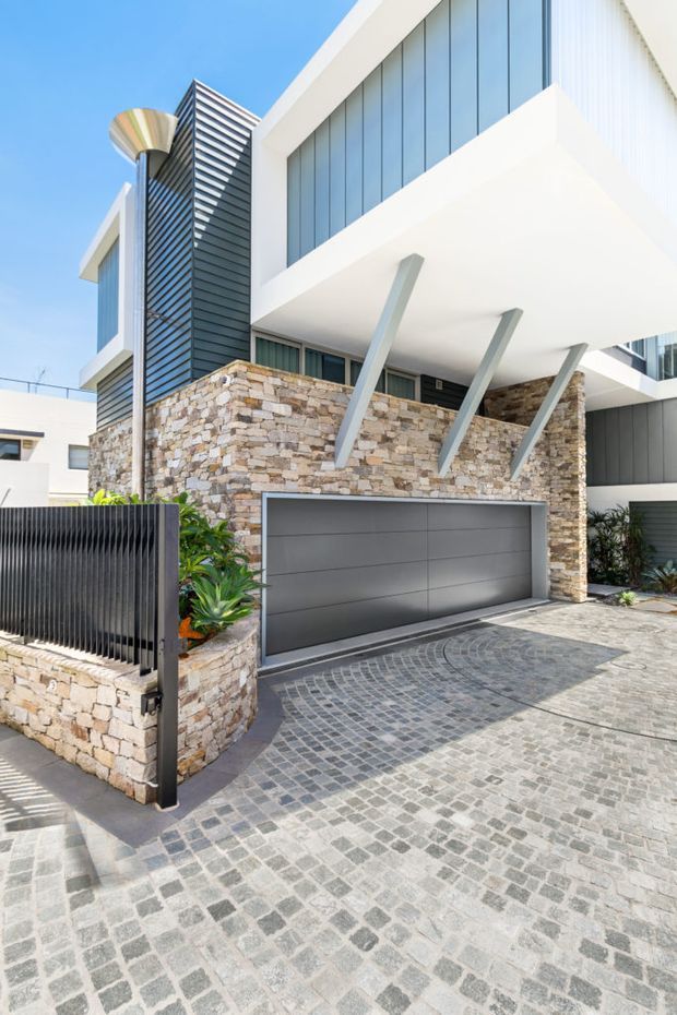 Street-level exterior view of a contemporary multi-storey residence. The facade combines stacked natural stone walls, white rendered projecting volumes, extensive blue-grey glazing, dark vertical metal cladding, and angled structural supports. A wide grey garage door opens onto a patterned grey cobblestone driveway, with a dark slatted boundary gate and landscaped planting at the entry.