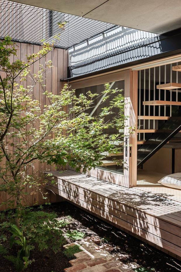 A light-filled courtyard beside a timber-clad home, with a leafy Japanese maple, raised timber decking, brick stepping path, and a partially enclosed stairwell. The stair features black steel stringers, timber treads, and vertical balustrade elements, while a patterned overhead screen casts strong shadows across the architecture.