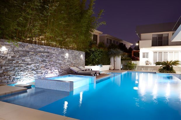 Night-time view of a landscaped residential swimming pool area with bright blue illuminated water, a raised spa or shallow pool section, stone retaining wall, bamboo planting, lounge chairs, and a contemporary white house in the background.