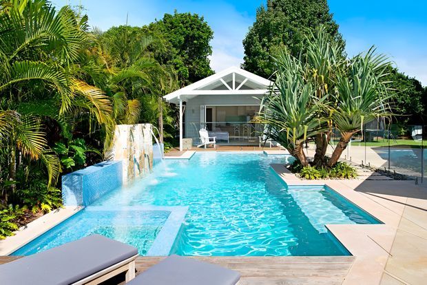A bright tropical outdoor pool area with a large turquoise swimming pool, integrated shallow ledges and water feature, surrounded by lush palms and architectural planting. A small light-coloured pavilion with a pitched gable roof and glass frontage sits beyond the pool, while paved decking and lounge furniture frame the foreground.