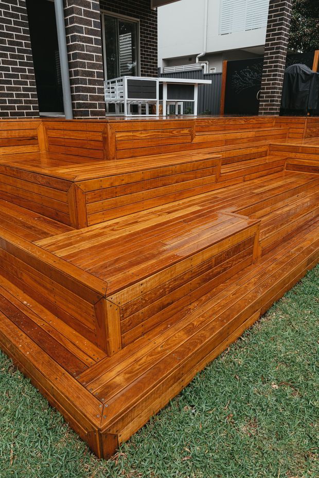 A multi-level outdoor timber deck and entertaining platform with broad built-in stepped seating or planter-like tiers. The deck is finished in warm, richly stained timber with visible board joints and fasteners. It sits beside a dark brick-and-render residential facade, with outdoor furniture, a barbecue cover, and lawn visible around it.
