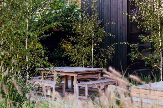 A landscaped outdoor dining area with a timber picnic-style table and benches, surrounded by ornamental grasses, shrubs, and slender trees. The space sits beside a dark vertical timber-clad building, with a raised timber deck or platform visible at right. The scene has a natural, secluded garden character.