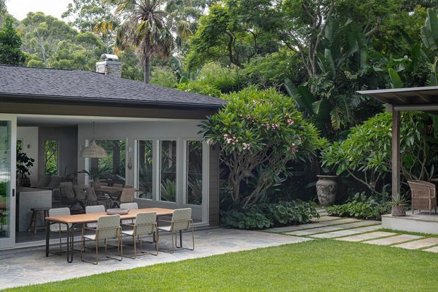 A single-storey contemporary house opens onto a lush tropical garden through extensive glazed doors. The covered outdoor living area includes a dining table and chairs, while stepping-stone paving crosses a manicured lawn toward a sheltered garden structure. Dark roofing, pale rendered walls, timber-toned outdoor furniture and dense greenery create a calm, resort-like setting.
