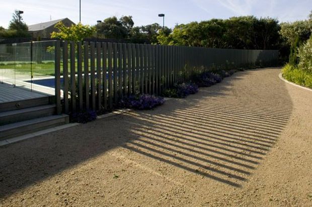 A landscaped outdoor walkway curves alongside a dark vertical-slat fence. Timber decking and clear glass pool or terrace balustrading appear at the left, with low purple planting along the fence and dense greenery beyond. Strong afternoon sunlight casts long striped shadows across the light gravel or compacted aggregate path.