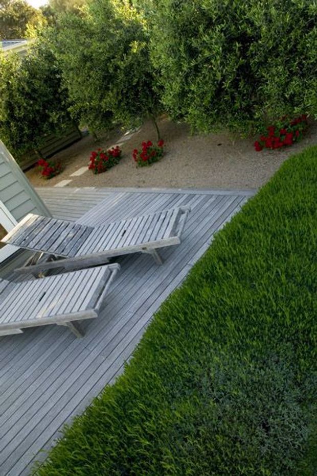 Elevated view of a landscaped outdoor terrace with a weathered grey timber deck, two matching reclining sun loungers, dense clipped evergreen hedging, gravel planting beds, and red flowering plants. The scene has a private, garden-focused residential character.