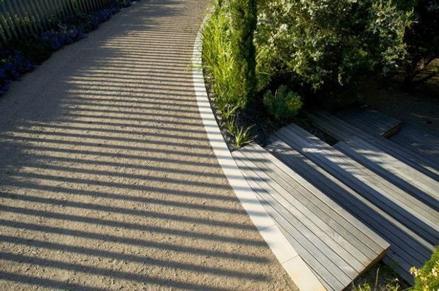 A landscaped outdoor pathway curves alongside a timber deck and stepped seating area. Strong linear shadows from a nearby fence fall across the pale gravel path, while dense planting borders the deck and walkway.