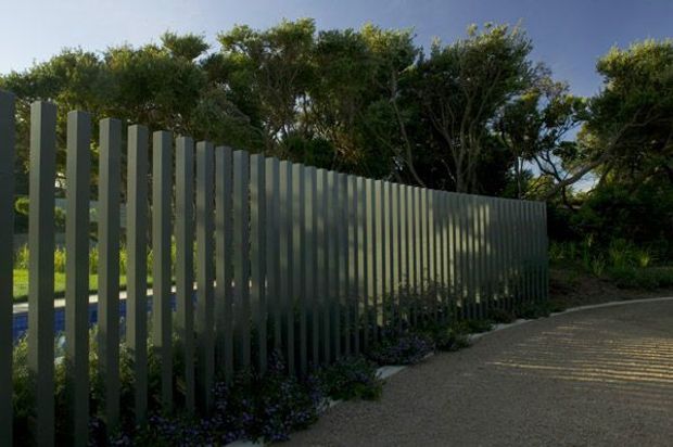Exterior view of a tall, regularly spaced vertical metal-slat screen or fence curving along a paved walkway. The muted grey-green slats create strong linear shadows, with dense planting and trees behind them and a glimpse of blue water or pool visible through the gaps.